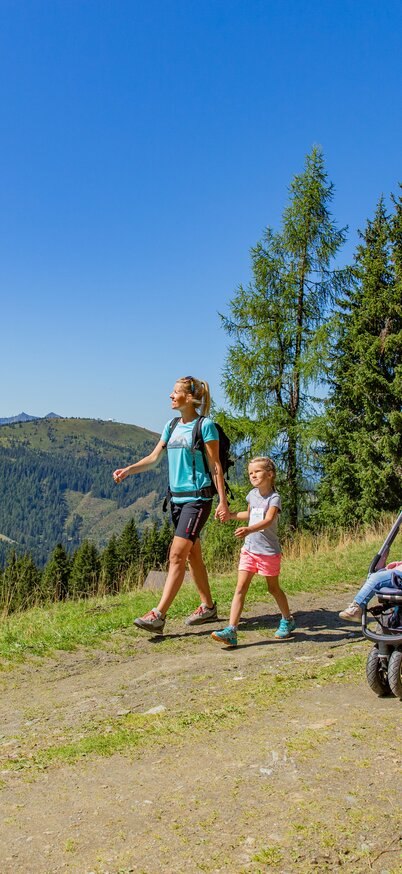 Eine Familie wandert mit Kinderwagen auf einem breiten Almweg am Hauser Kaibling, umgeben von Bergen und Wäldern. | © René Eduard Perhab