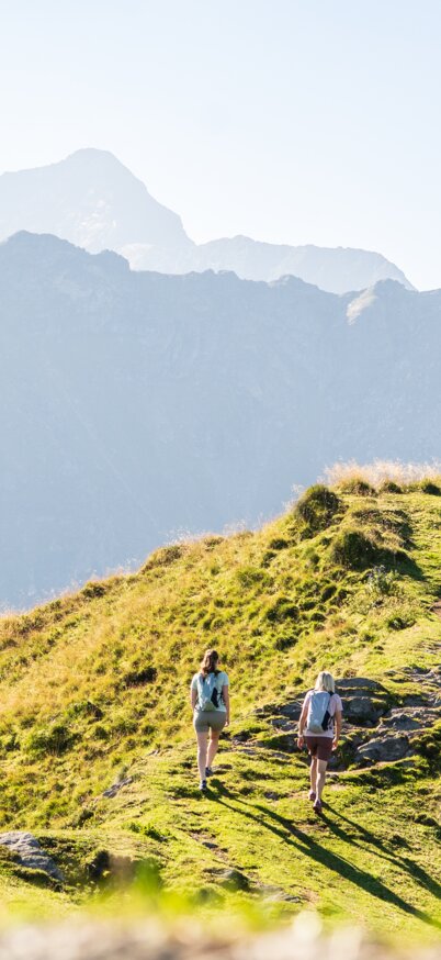 Zwei Wanderinnen gehen über einen grünen Höhenrücken mit weitem Blick auf die Berglandschaft der Schladminger Tauern im Sonnenlicht. | © Josh Absenger