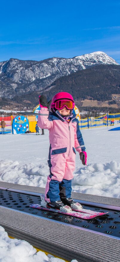 Kinder fahren auf dem pinken „Weidas Zauberteppich“ im Wollis Kids Park am Hauser Kaibling den Förderbandlift hinauf. Ein Mädchen im rosa Skianzug winkt lachend in die Kamera, während hinter ihr weitere Kinder mit Ski und Laufrädern begleitet von einer Erwachsenen den Zauberteppich nutzen. | © René Eduard Perhab