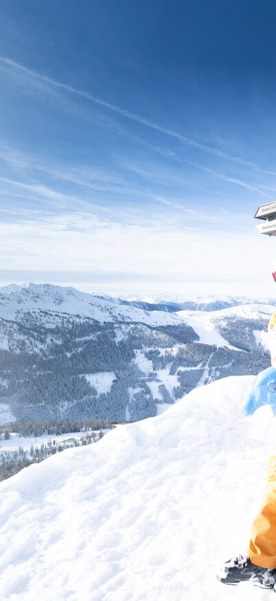 Two female skiers laughing at the summit cross of the Hauser Kaibling, skis in their arms, surrounded by a panorama of snow-covered mountains under a blue sky. | © Bernhard Moser
