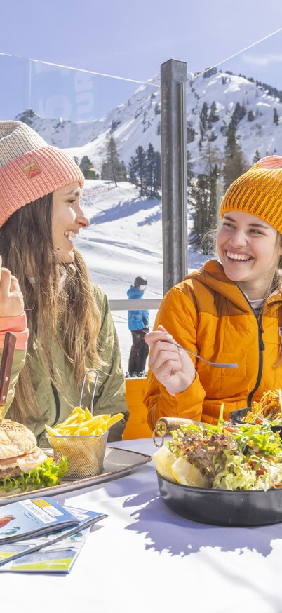 Zwei Frauen essen Burger, Pommes und Salat auf der Sonnenterrasse der Kaiblingalm am Hauser Kaibling, im Hintergrund die Skipiste und verschneite Gipfel. | © Bernhard Moser