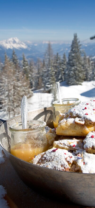 Fresh Kaiserschmarrn with powdered sugar and apple sauce served in a pan in the snow, with a view of the slopes and mountains at Hauser Kaibling. Behind it, an old pair of wooden skis. | © Herbert Raffalt